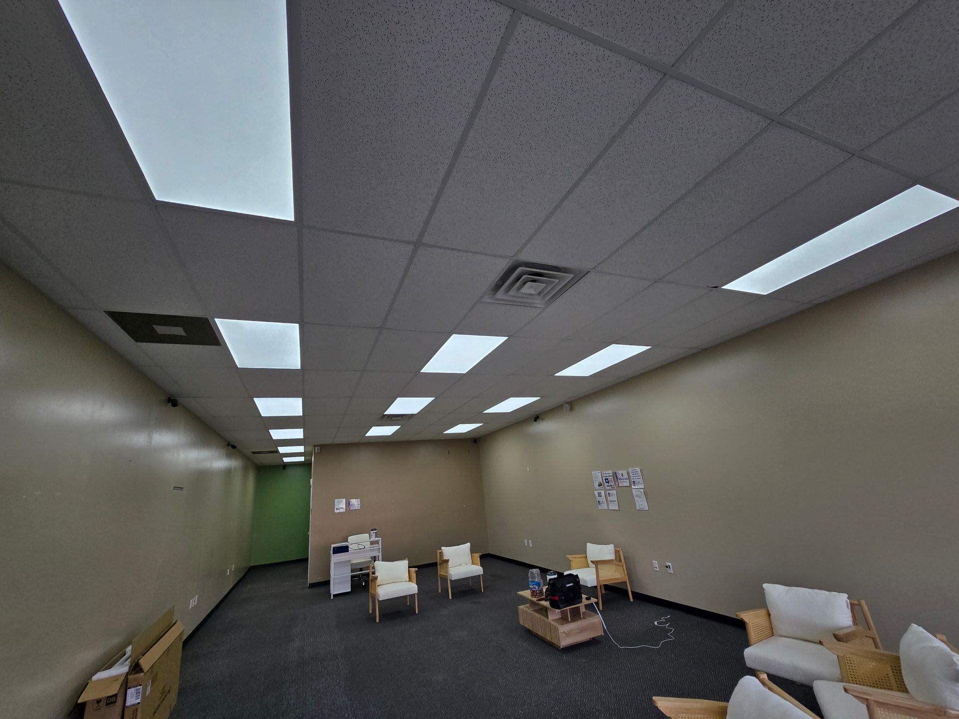 An empty waiting room with beige walls, a dark carpet, and several chairs. Fluorescent lights are in the ceiling.