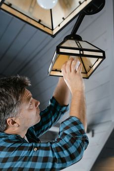 Man changes lightbulb in a craftsman-style fixture; he's wearing blue plaid and is on a wooden ceiling.