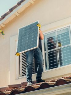 Solar panel installer carries a solar panel on a rooftop in front of a window.