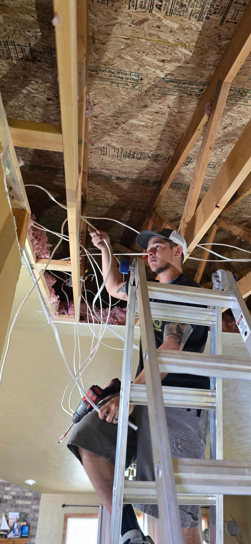 A man on a ladder working on electrical wiring in an unfinished ceiling.