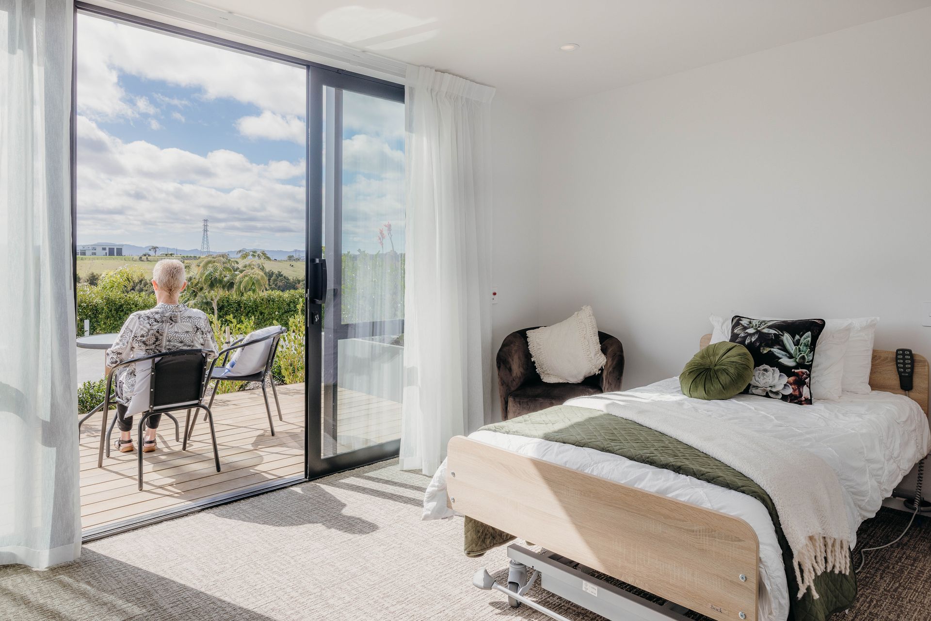 A person sits on a balcony overlooking a scenic view. A bedroom with a bed and armchair.