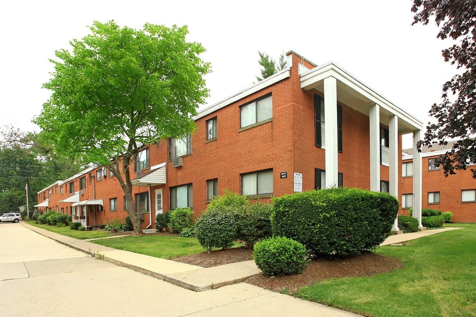 Two-story brick apartment buildings with white columns, landscaping, and a street.