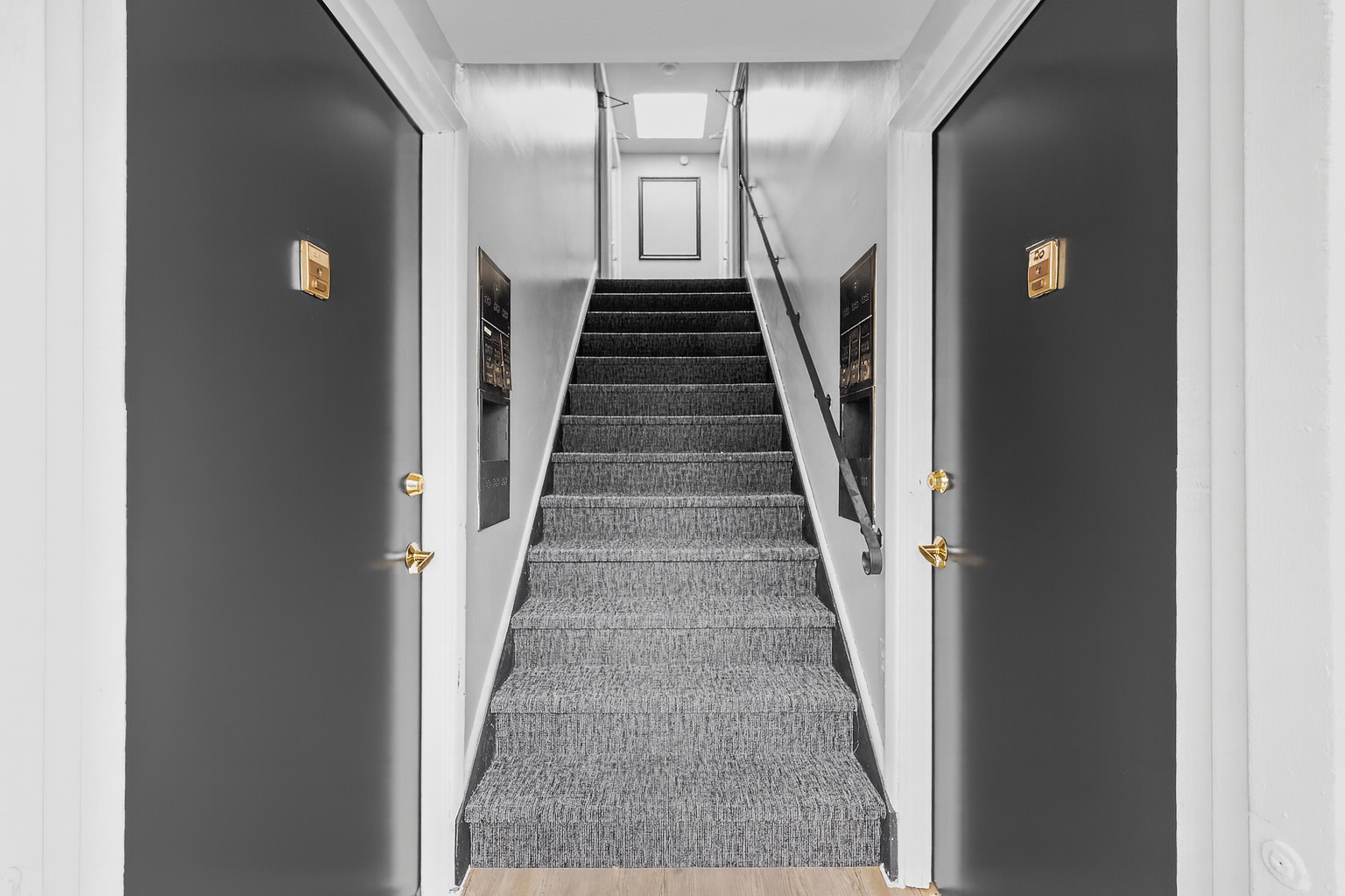 Narrow hallway with stairs, two dark gray doors on each side, and a skylight at the top of the stairs.