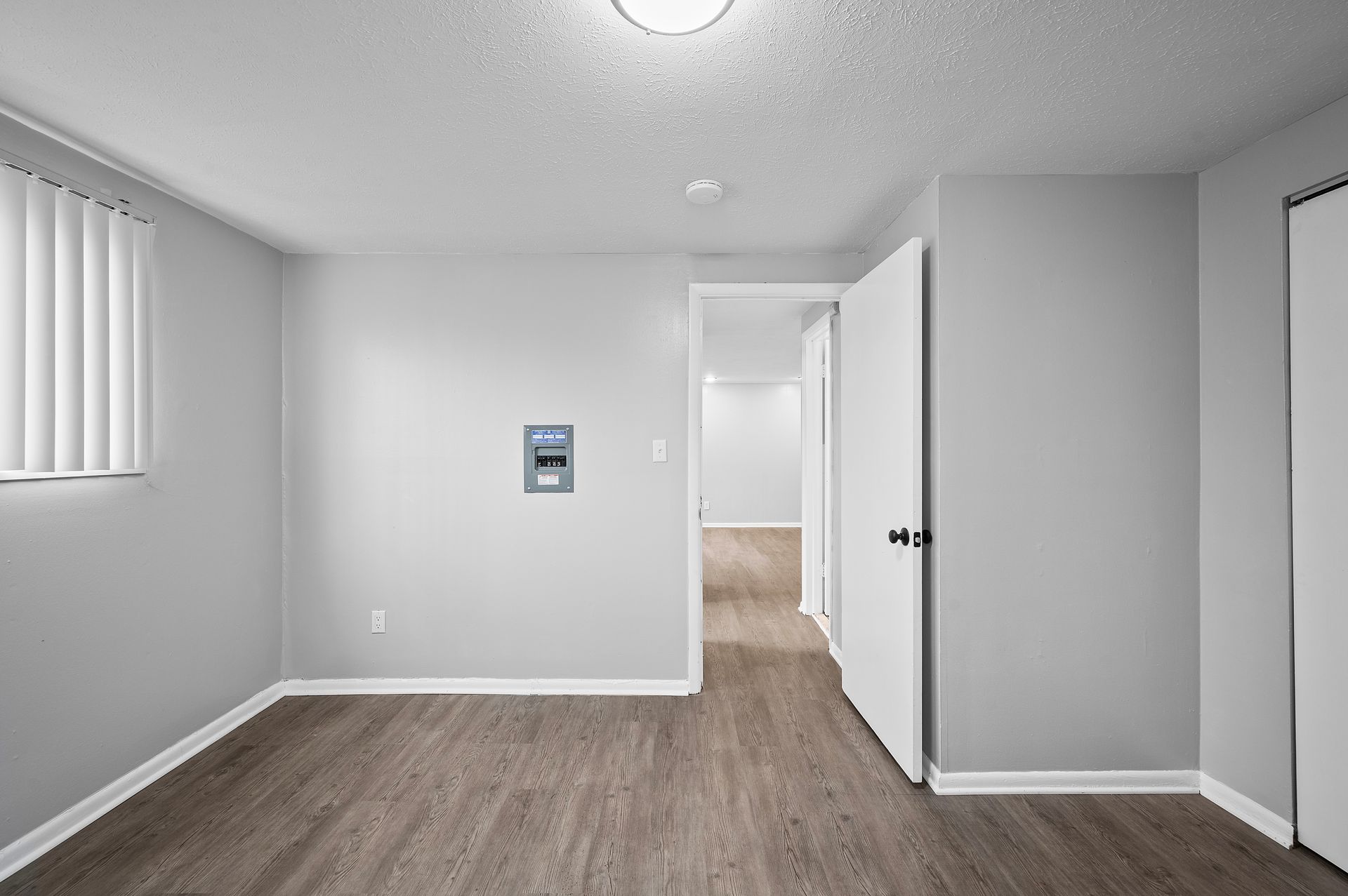 Empty room with light gray walls, wood-look flooring, white door, and window with vertical blinds.