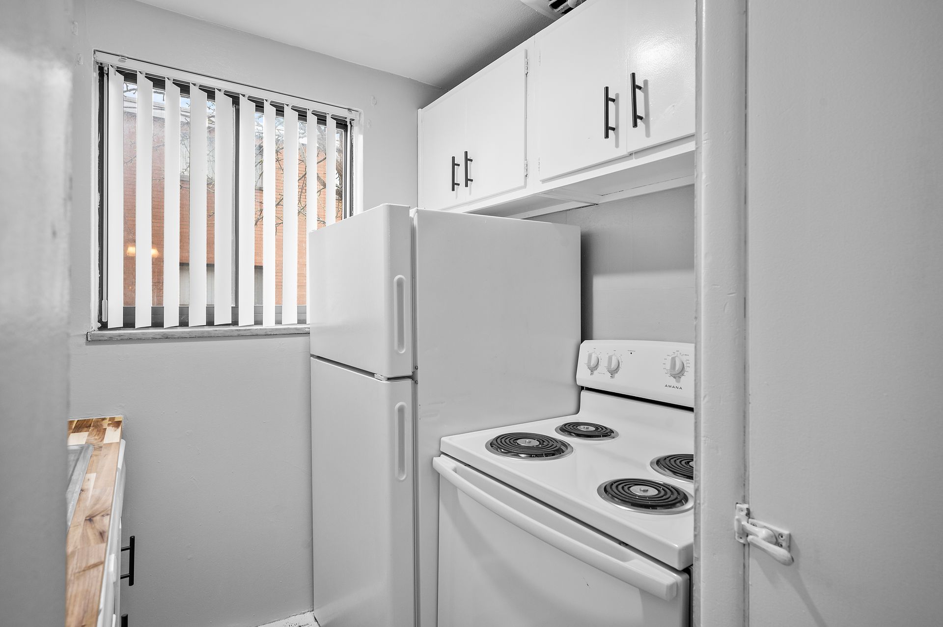 Small white kitchen with fridge, stove, cabinets, and window with blinds.
