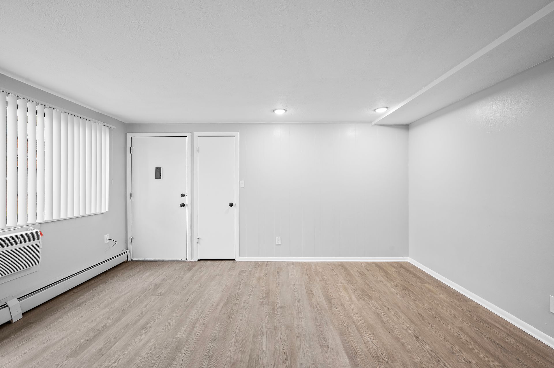 Empty room with pale wood flooring, white walls, and a window with blinds.