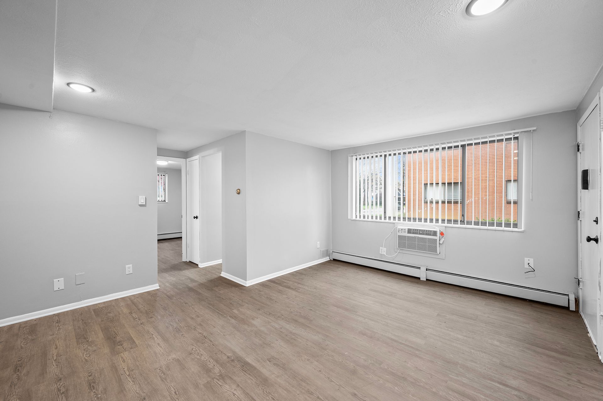 Empty, brightly lit apartment room with gray walls, wood-look flooring, and a window with a view of a brick building.