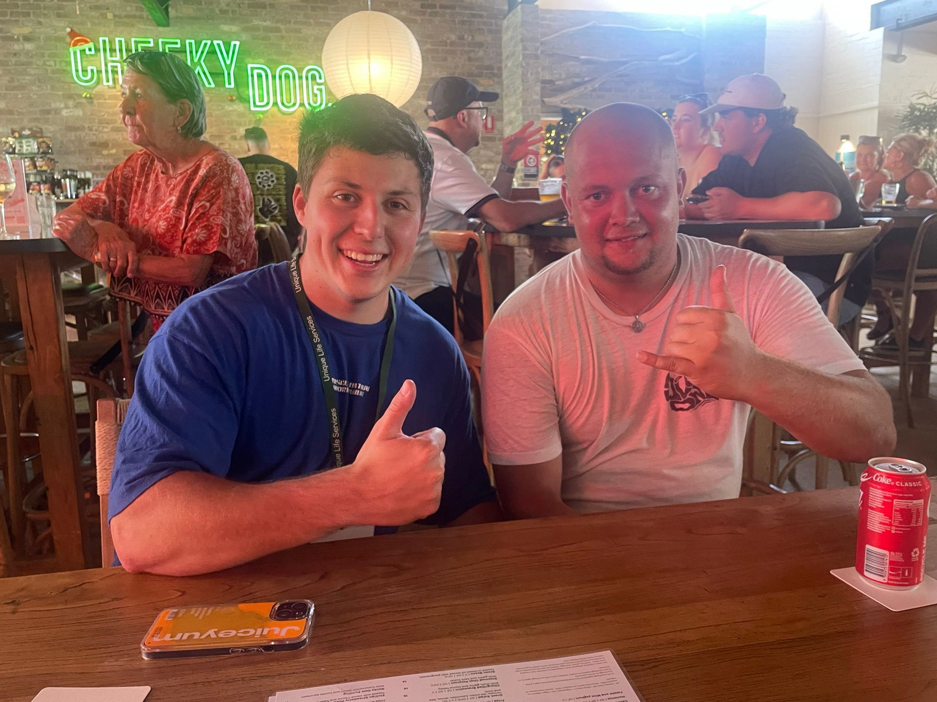 Two men at a bar giving thumbs-up. One in blue shirt, other in white. 