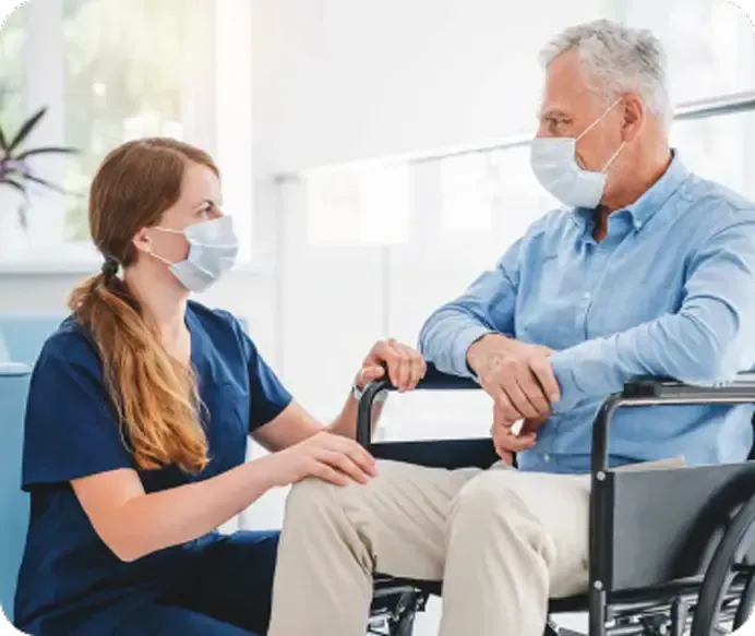 Nurse in scrubs comforting a person in a wheelchair, both wearing face masks, in a medical setting.