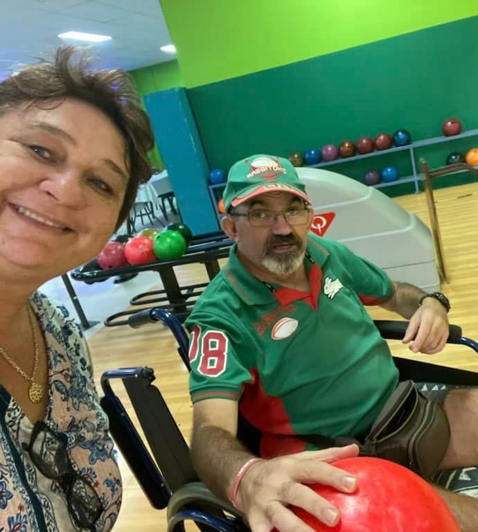 Woman taking a selfie with a man in a wheelchair holding a bowling ball, inside a bowling alley.