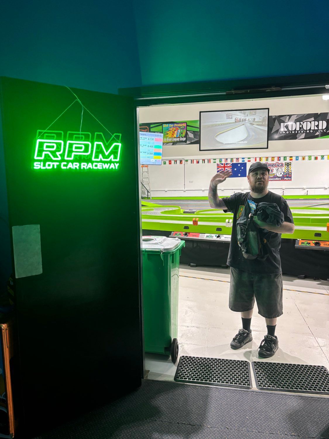 Man waves from the doorway of RPM Slot Car Raceway, under green neon sign. The racing track is visible in the background.