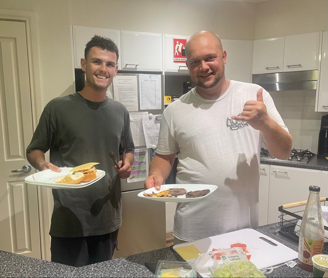Two men in a kitchen holding plates of food, smiling. One gives a thumbs up.