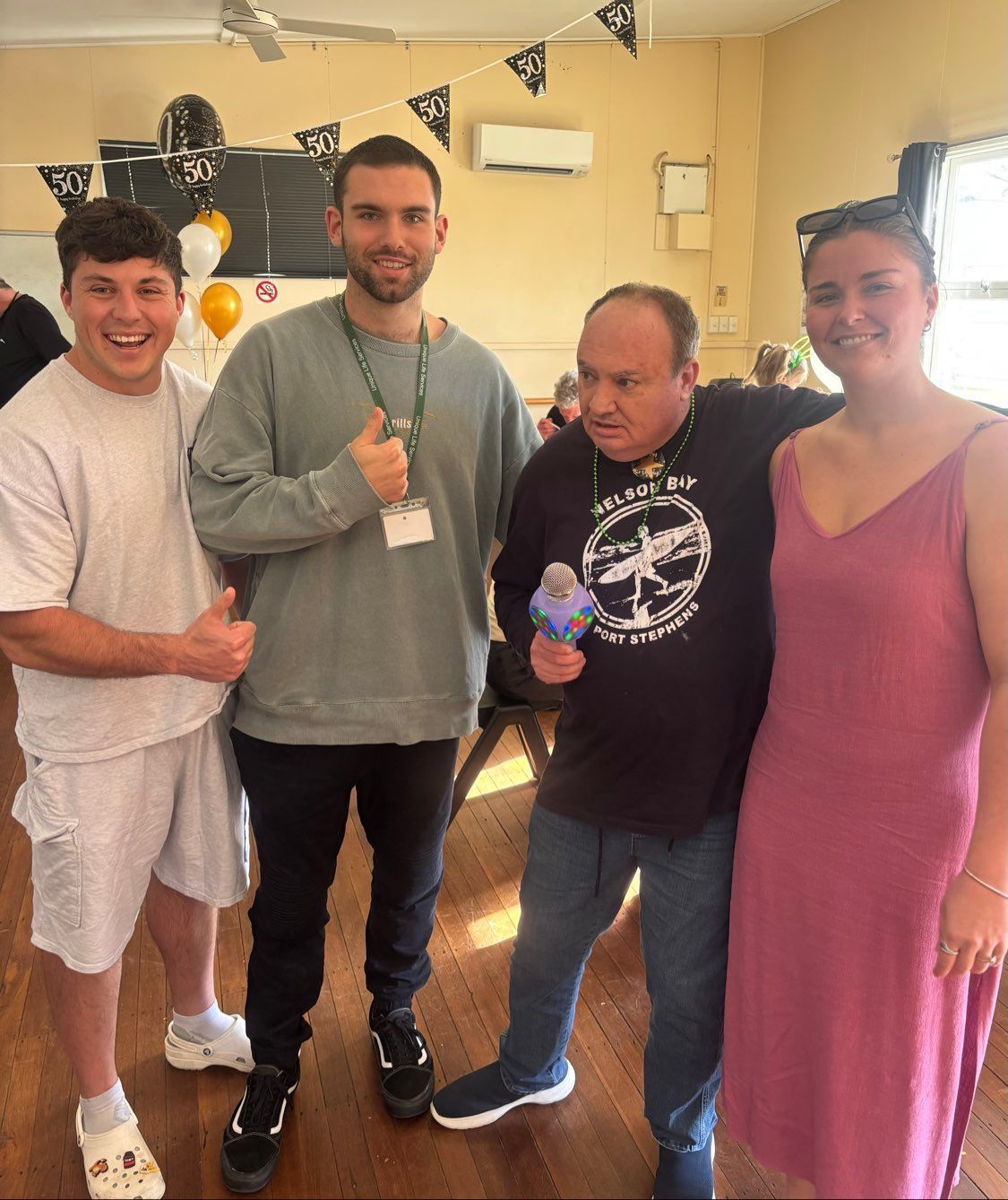Four people smiling and posing indoors. A man holds a microphone. Balloons and decorations visible.