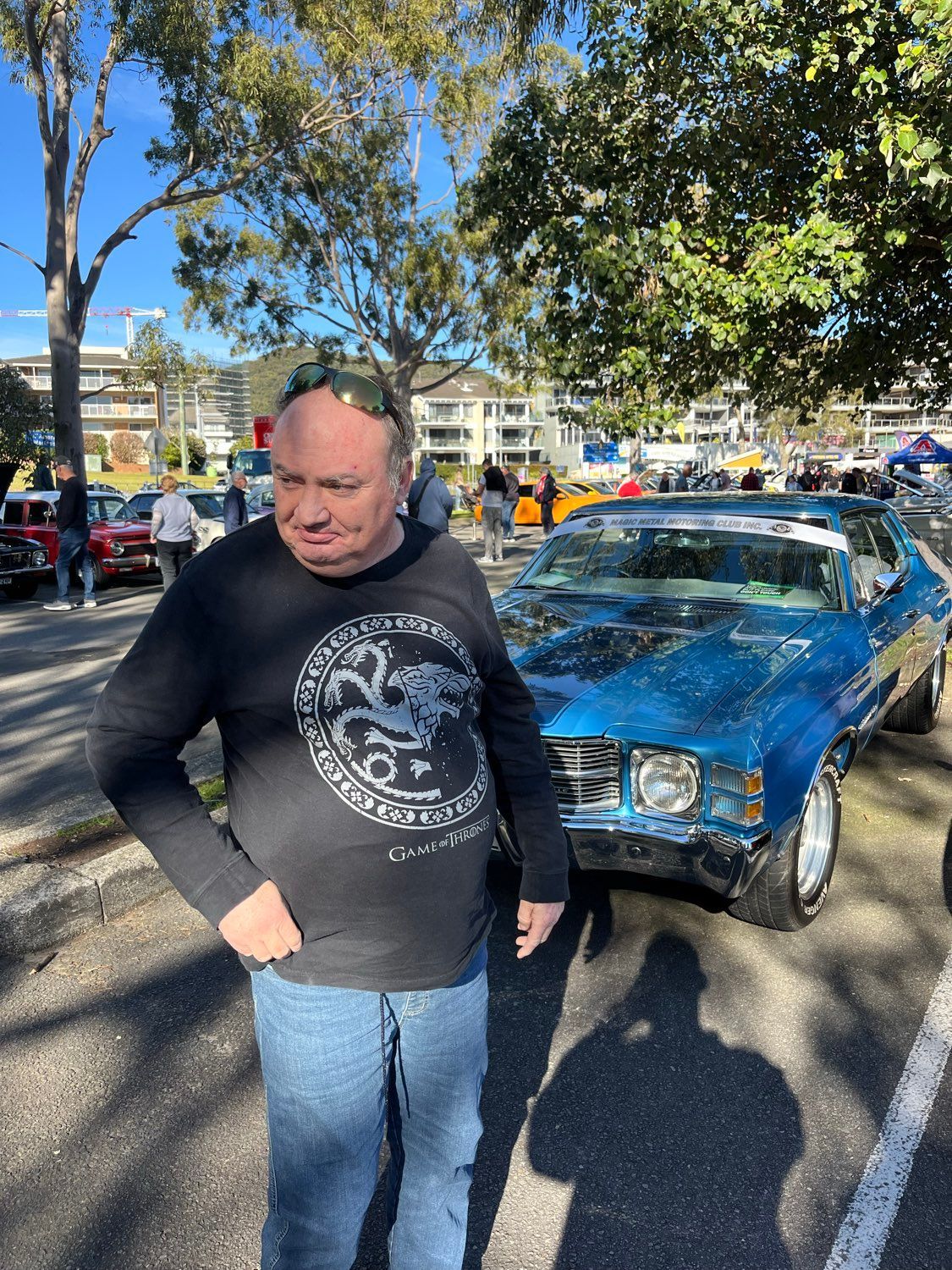 Man in black shirt stands next to a blue classic car. Outdoors, sunny day.