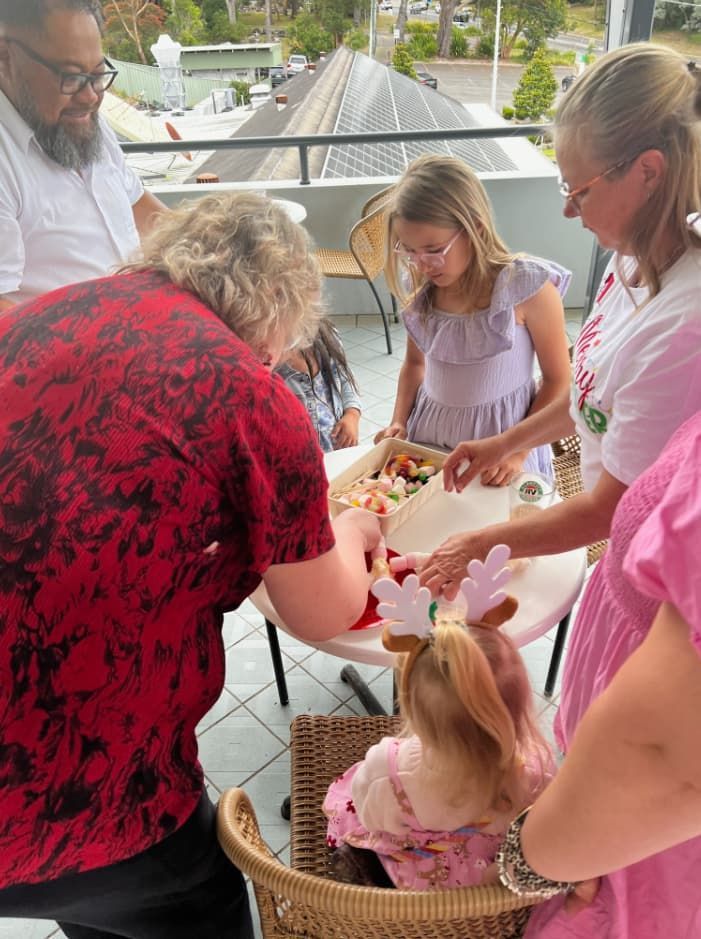 A Group Of People Are Standing Around A Table With A Little Girl Sitting In A Chair — Unique Life Services In Nelson Bay, NSW