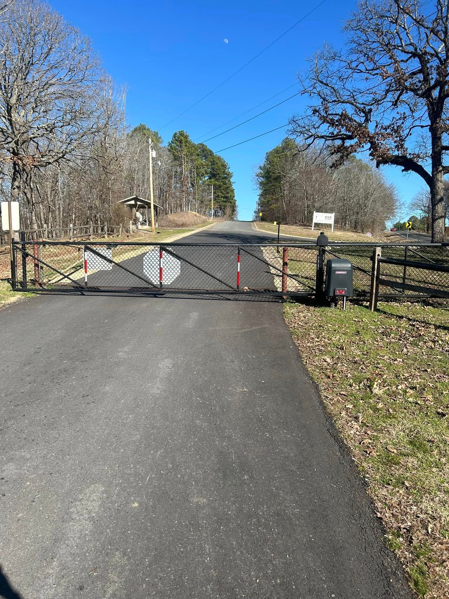 Gated asphalt driveway leading uphill; trees on either side and clear blue sky.