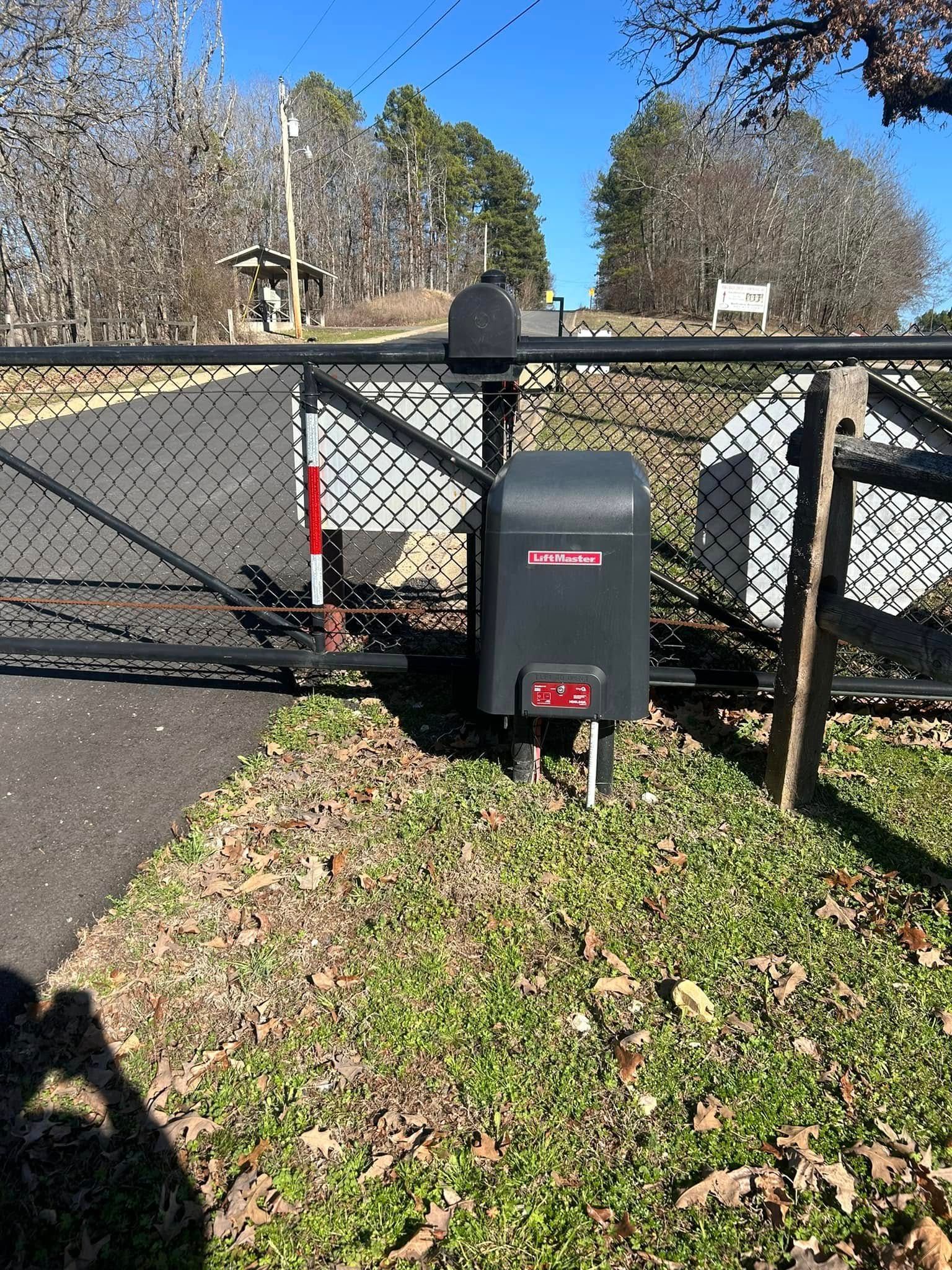 Black gate with automatic opener, mailbox, and chain link fence in front of a driveway, in a grassy area.