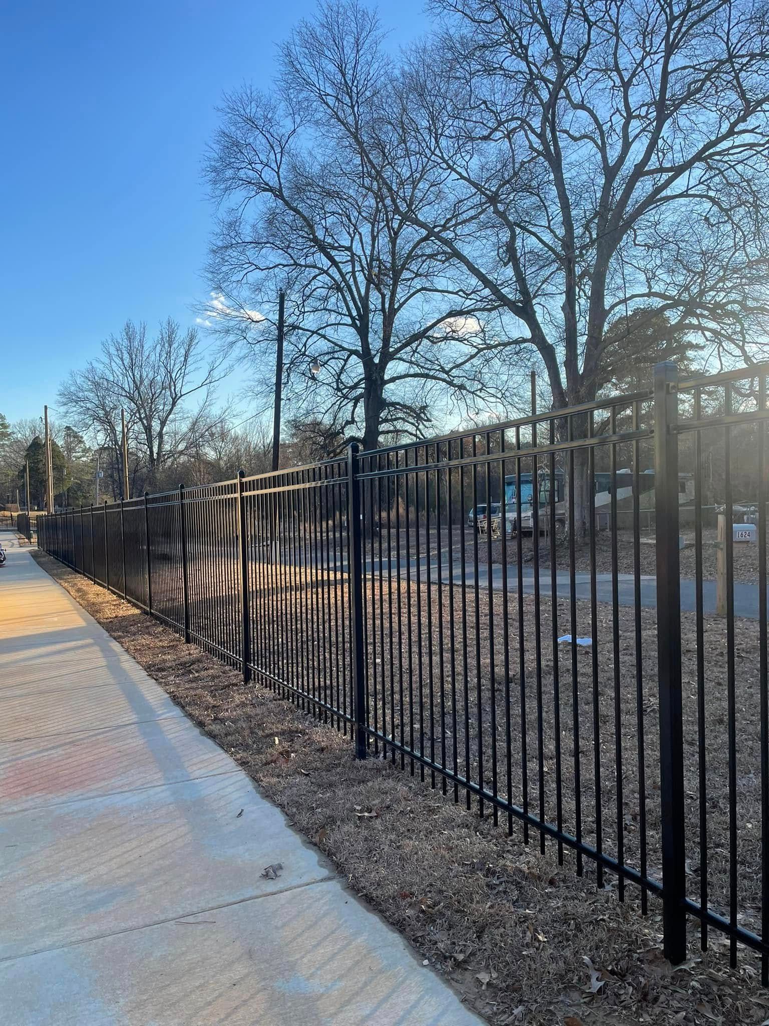Black metal fence alongside a sidewalk; bare trees in the background under a blue sky.