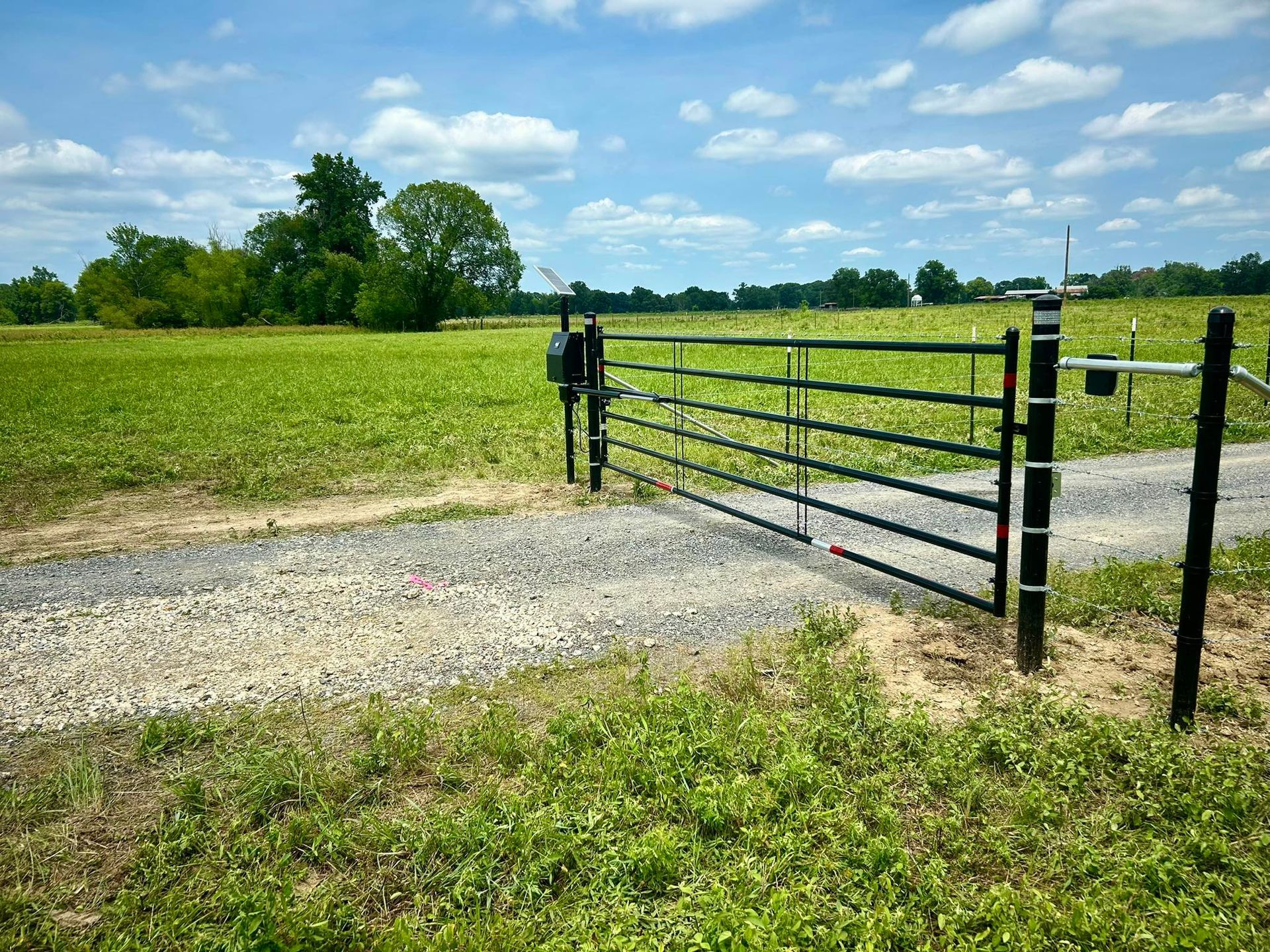 Black metal gate on a gravel drive leading into a green field, under a cloudy blue sky.