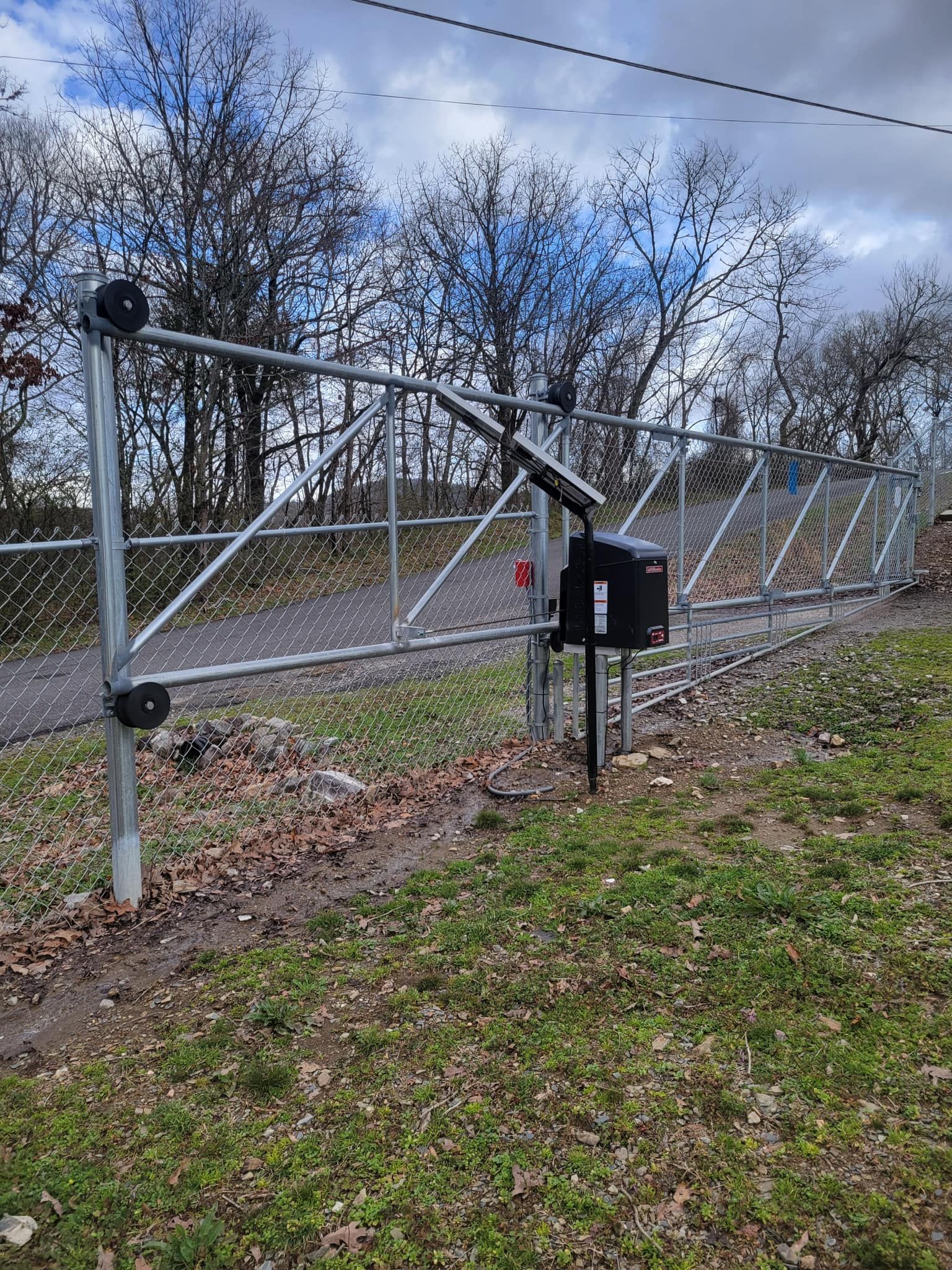 A chain-link fence gate, opened by an automated arm. It is outdoors on a grassy verge, with trees in the background.
