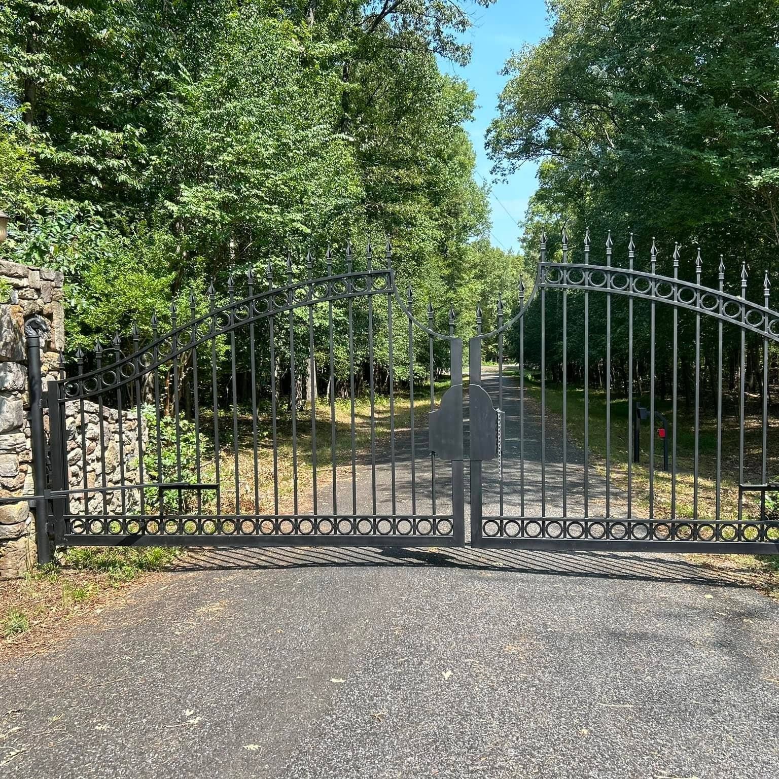 Black metal driveway gate, open, leads to a tree-lined driveway, on a sunny day.
