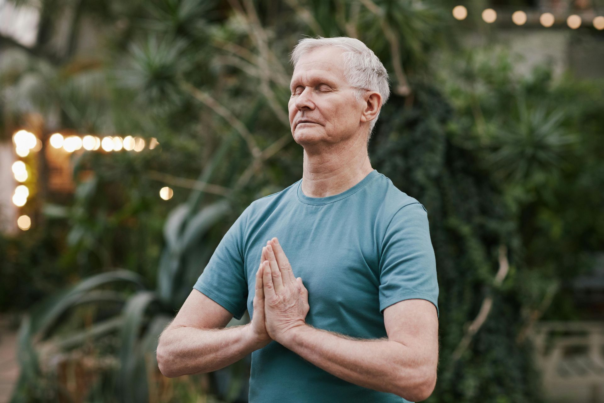 Man with closed eyes, hands together in prayer, outdoors with greenery and lights.