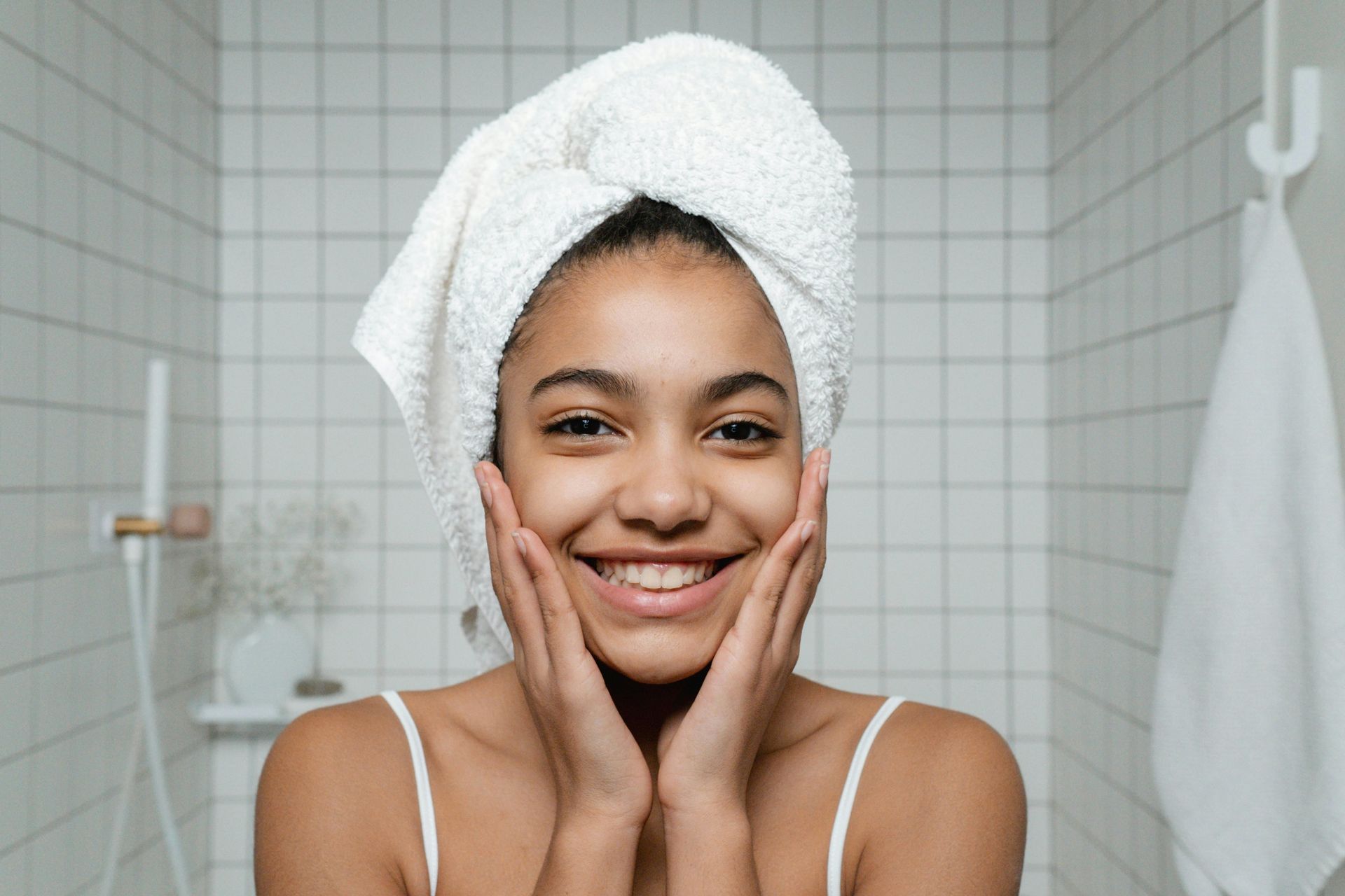 Woman with towel on head smiles, hands on cheeks, in a white-tiled bathroom.