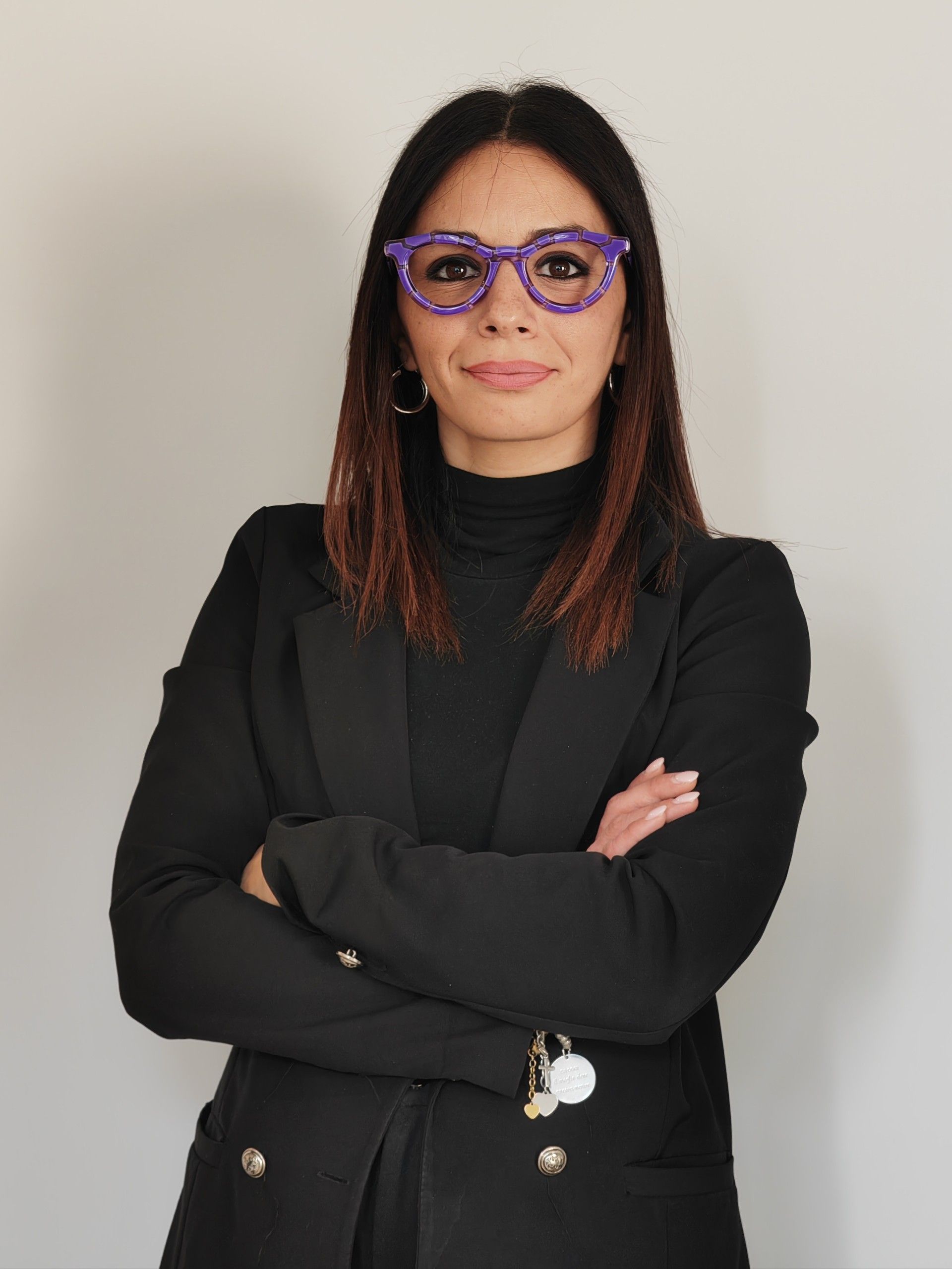 Una donna che indossa occhiali viola e una giacca nera è in piedi con le braccia incrociate.