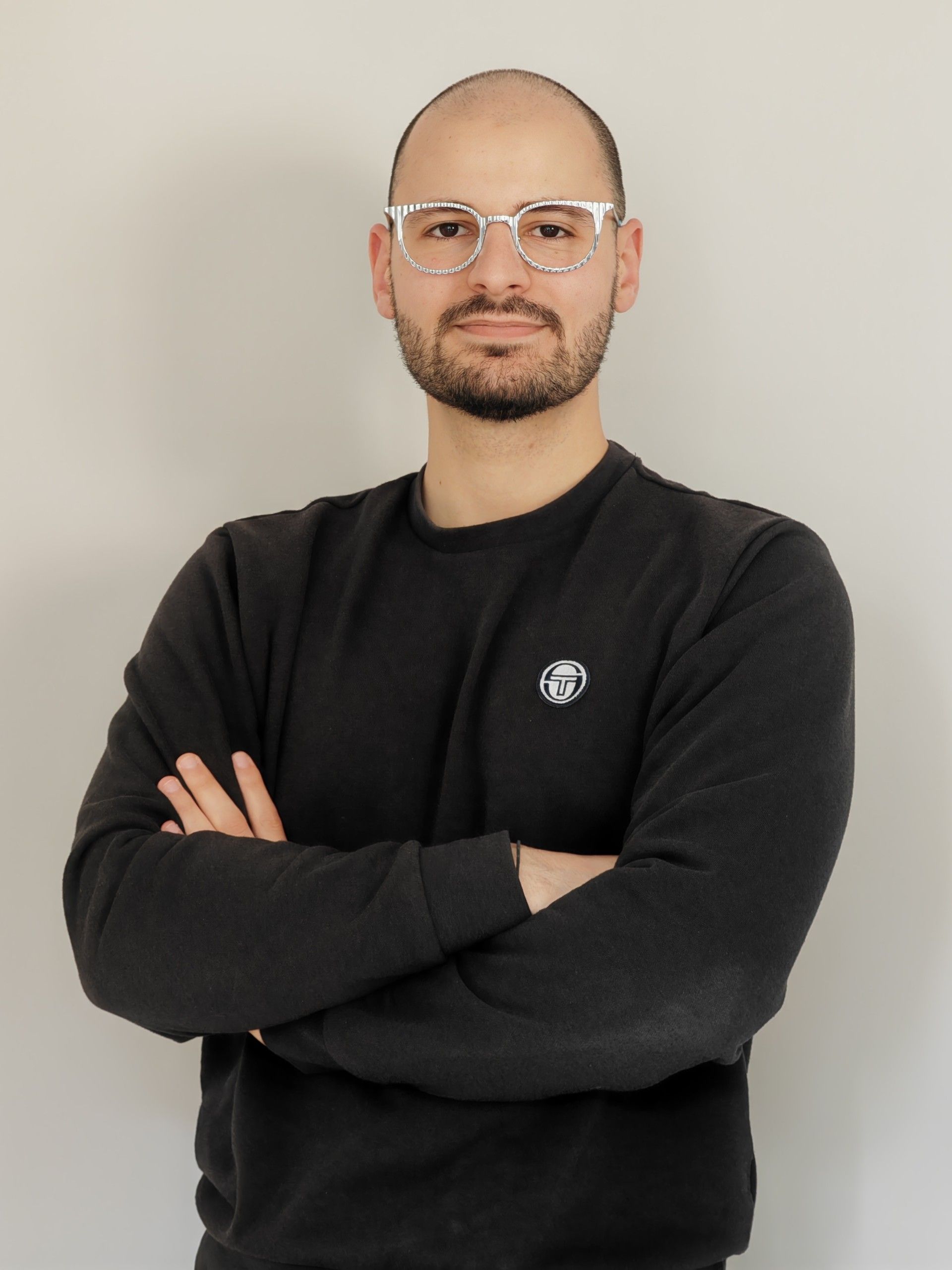 Un uomo con gli occhiali e un maglione nero è in piedi con le braccia incrociate.