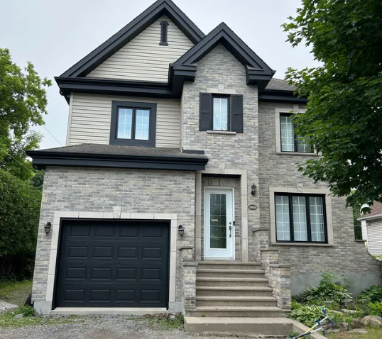 Maison à deux étages avec façade en pierre, boiseries noires, porte de garage et marches d'entrée.