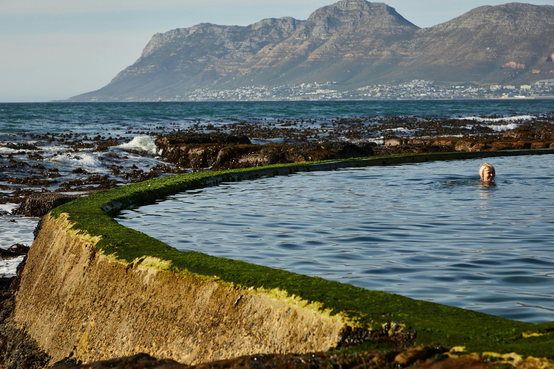 A man is swimming in a pool of water with mountains in the background.