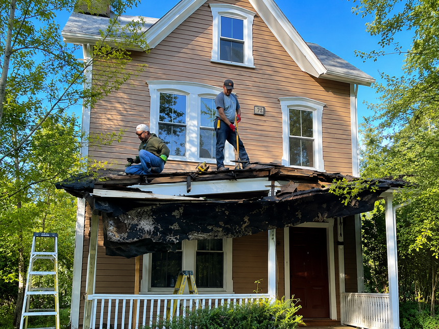 Workers repairing a fire-damaged porch on a brick house under a blue sky