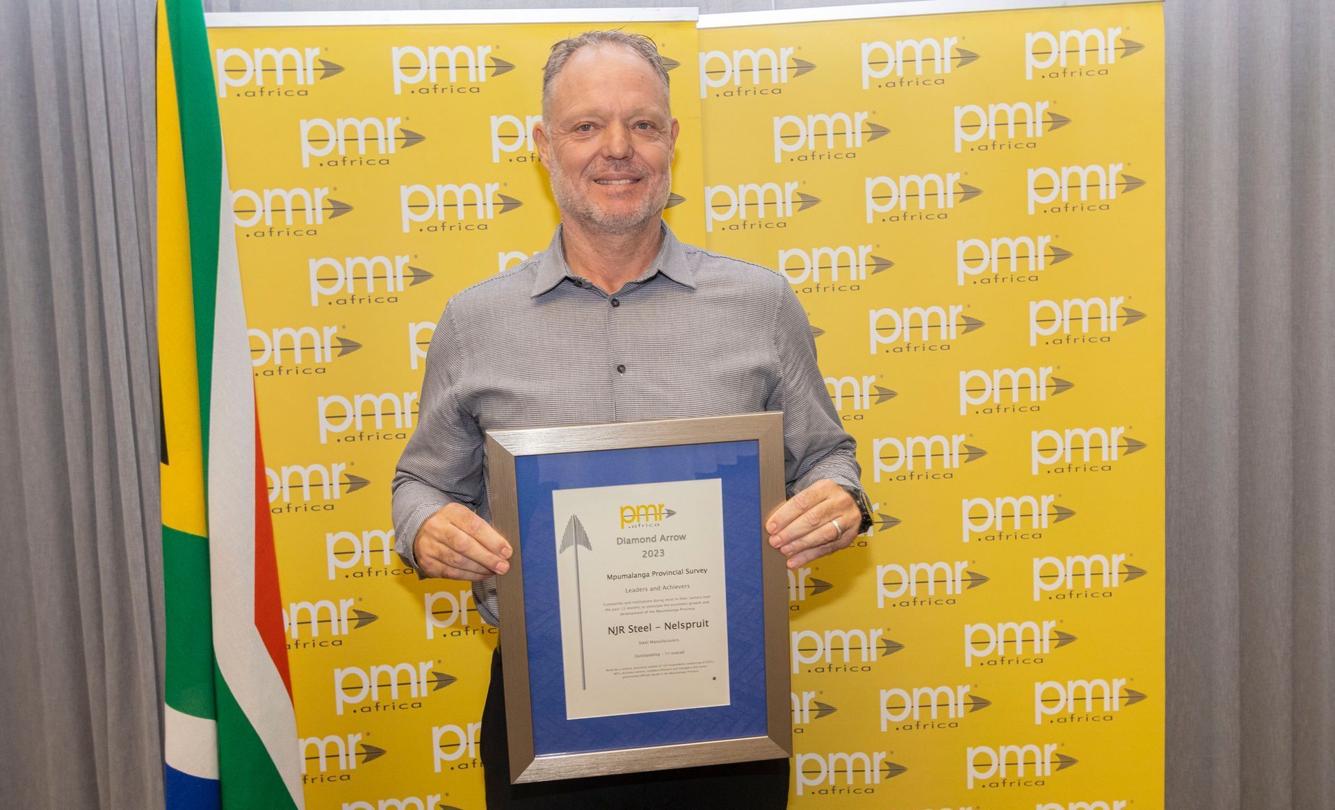 Andries de Kock, the Branch Manager of NJR Steel is holding a certificate in front of a yellow wall.