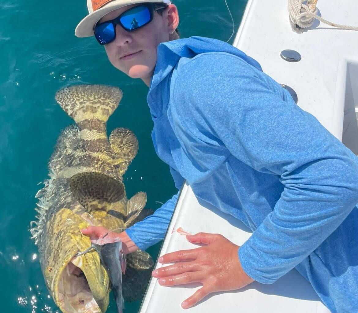 A man wearing sunglasses and a hat is holding a large fish on a boat.