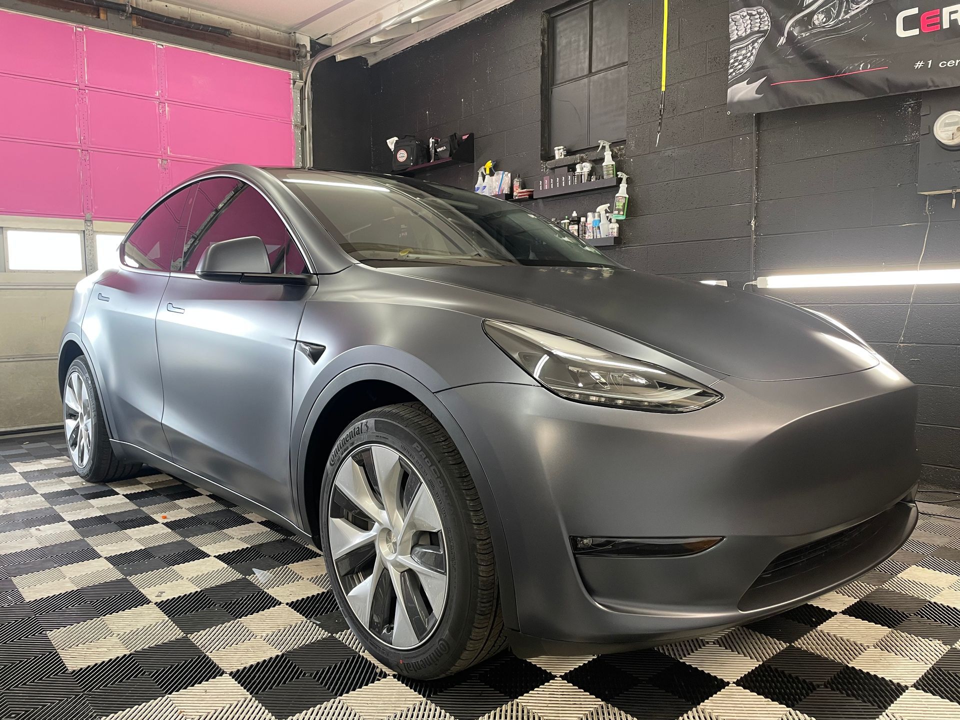Gray Tesla Model Y car in a garage with a checkered floor, tinted windows, and matte finish.
