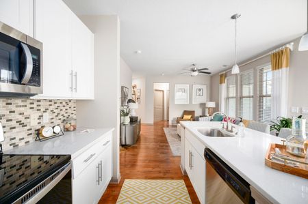 Modern white kitchen with stainless steel appliances, marble countertops, and a view into a living area with hardwood floors.