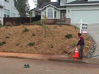 A man is cleaning the sidewalk in front of a house with a for sale sign in the background.