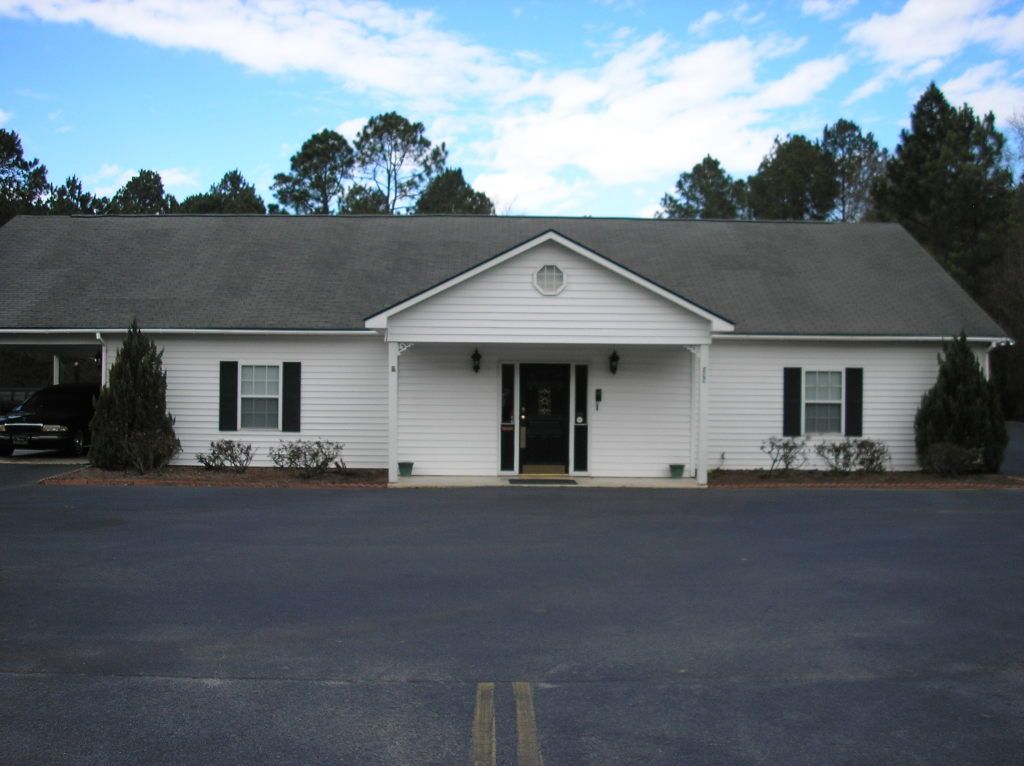 Small white building with a front porch and dark roof, seen across an empty parking lot