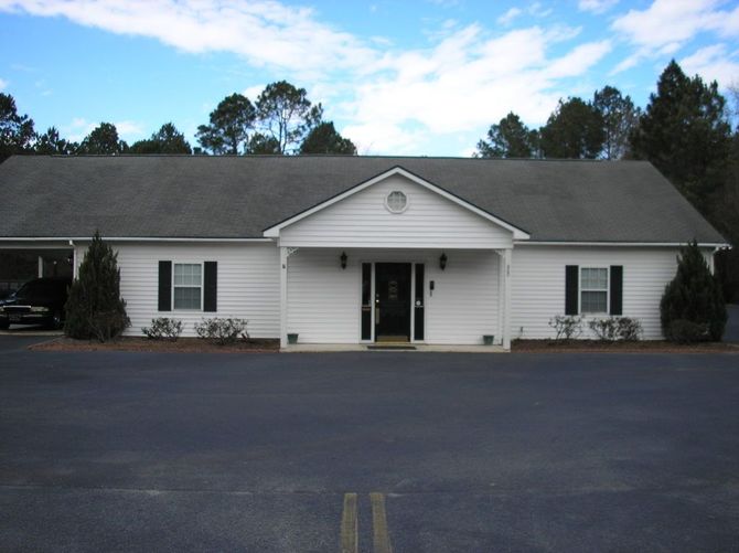 Small white building with a front porch and dark roof, seen across an empty parking lot