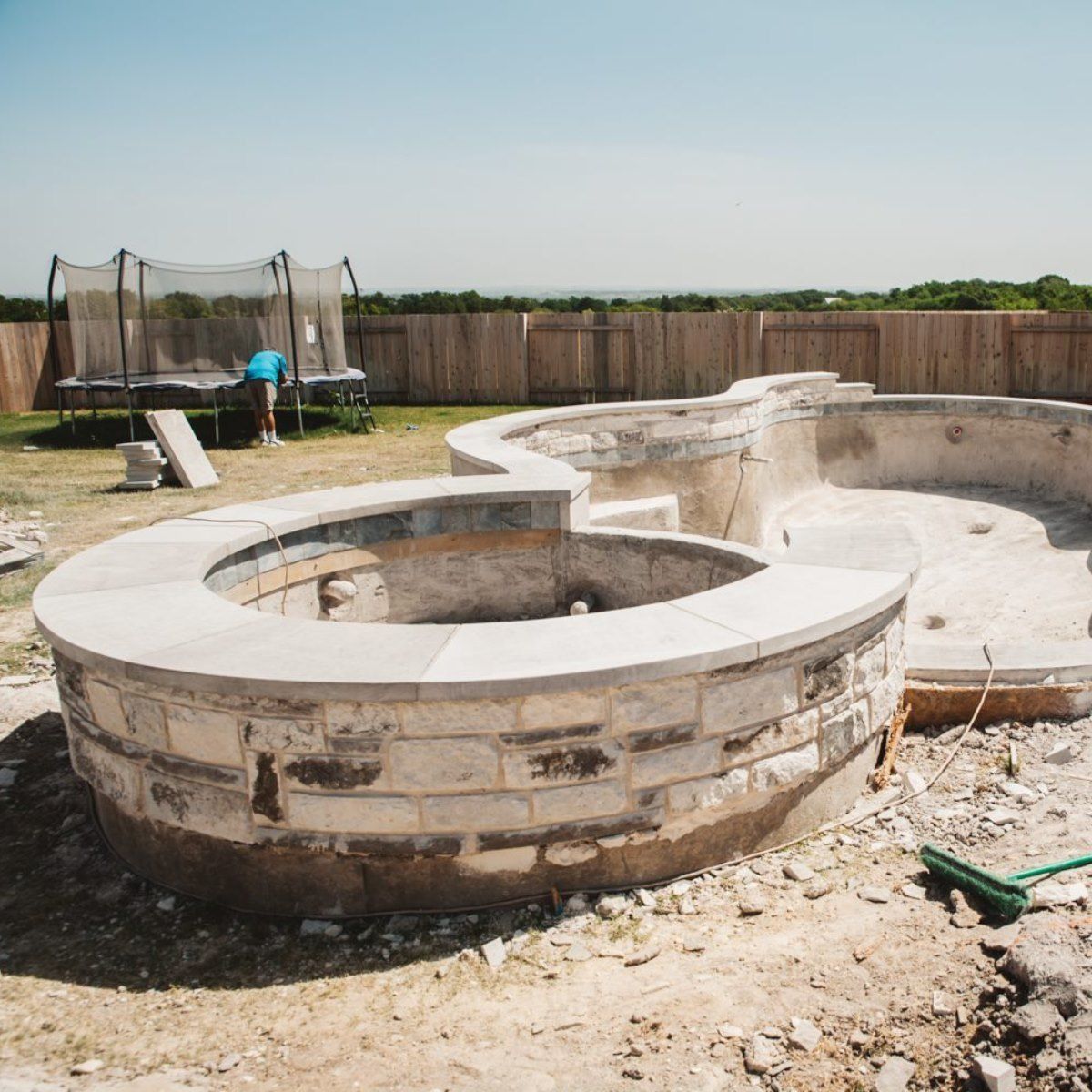 Stone-walled pool under construction in backyard with a person near a trampoline. Sunny day.