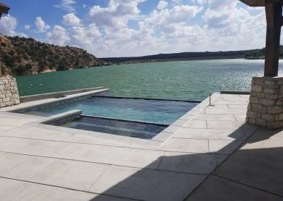 A pool overlooking a lake under a partly cloudy sky. Stone pillars and patio surround the pool.