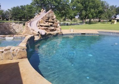 Pool with a rock slide in a grassy yard on a sunny day.