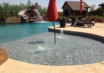 Pool with a slide, waterfall, and shallow area with fountains, surrounded by a deck and an umbrella.