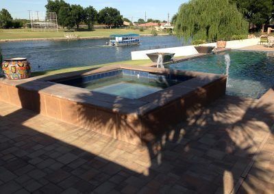 Hot tub and pool overlooking a lake. Sunny day, clear water, landscaping visible.