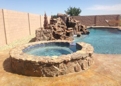 Pool with stone-lined hot tub and waterfall feature; adjacent to a swimming pool and tan walls under a blue sky.