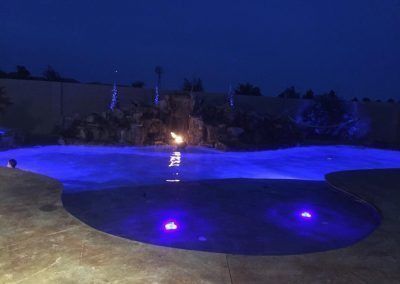 Nighttime view of a pool illuminated with blue lights, featuring a waterfall and decorative lights.