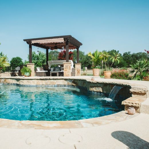 A pool with a waterfall feature and a pergola with seating on a sunny day.