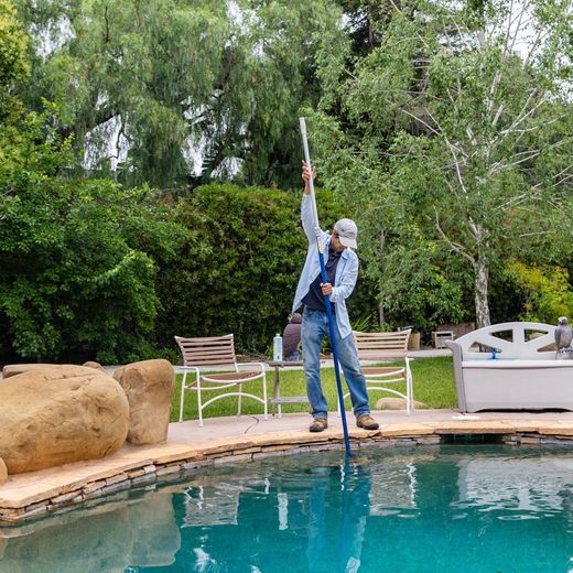 Man using a pool net, outdoors near a pool. He wears a hat, jeans, and a blue shirt.