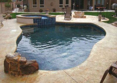 Swimming pool with hot tub, surrounded by concrete patio and landscaping in a backyard.