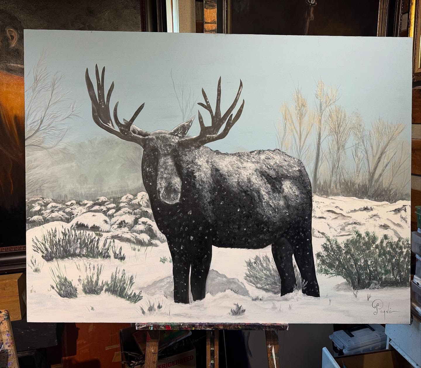 A painting of a moose standing in the snow on an easel.